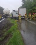 Samochód ciężarowy z cysterną po zderzeniu z naczepą drugiego samochodu ciężarowego, po lewej dwie karetki pogotowia, przed samochodami grupa dziewięciu strażaków.