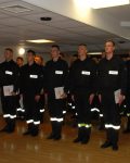 Grupa strażaków stojąca na baczność podczas wręczania dyplomów ukończenia kursu podstawowego, w dłoniach dyplomy