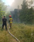 Budynek w lesie, jeden strażak na drabinie kieruje prąd gaśniczy na dach, przy wężu tłocznym dwóch strażaków i dwóch policjantów