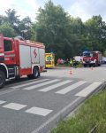 Wóz strażacki stoi na przejściu dla pieszych, za nim drugi wóz strażacki, karetka pogotowia i radiowóz policji. Pomiędzy nimi grupa Strażaków, cywili i jeden policjant.