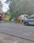 Na pierwszym planie samochód policji i wóz strażacki odgrodzony pachołkami, pomiędzy nimi trzech strażaków, ratownik medyczny i cywil, w tle dwa uszkodzone samochody poza pasem ruchu.