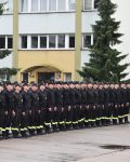 Grupa strażaków stoi na baczność w dwuszeregu, na tle wejścia głównego Szkoły.