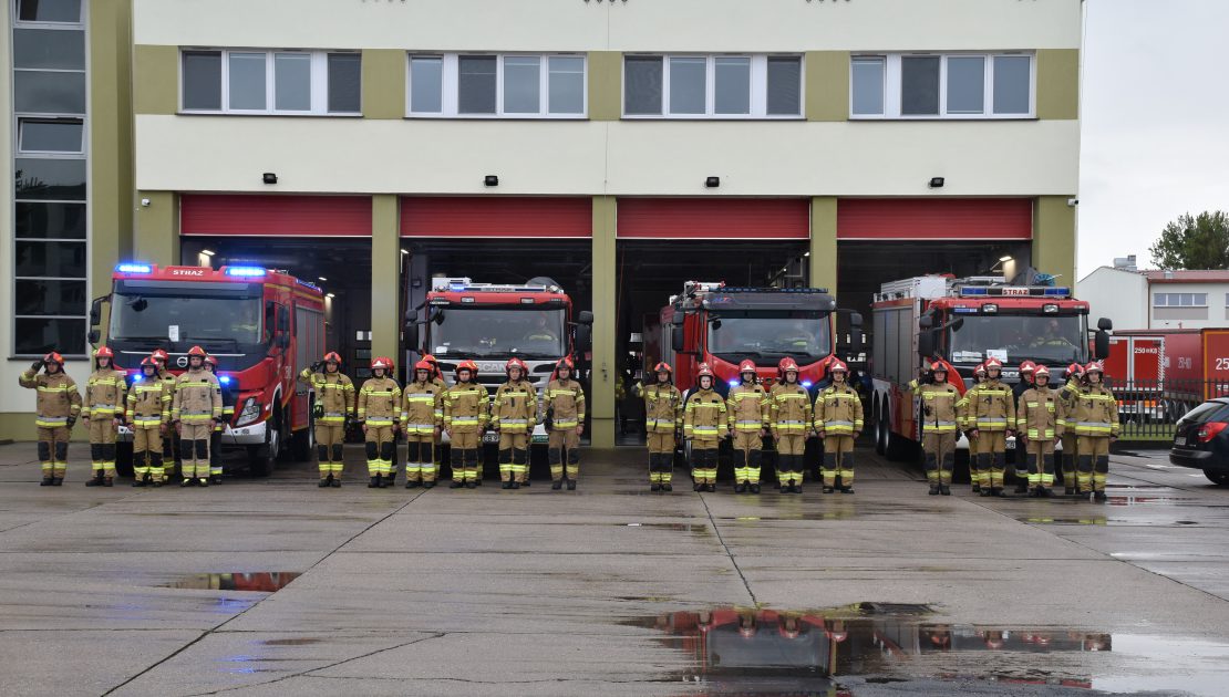 Cztery zastępy strażaków stoją na baczność przed wozami strażackimi, na tle garażu bojowego Szkoły, dowódcy zastępów oddają honor