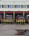 Cztery zastępy strażaków stoją na baczność przed wozami strażackimi, na tle garażu bojowego Szkoły, dowódcy zastępów oddają honor
