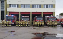 Cztery zastępy strażaków stoją na baczność przed wozami strażackimi, na tle garażu bojowego Szkoły, dowódcy zastępów oddają honor