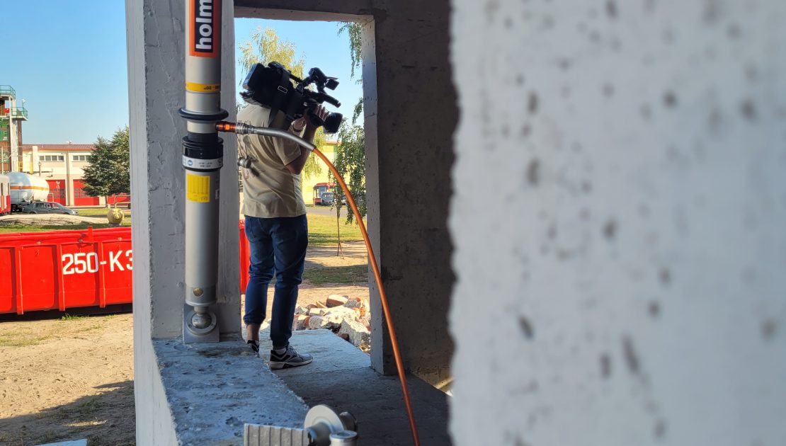 Budynek szkoleniowy, z zamontowaną hydrauliczną podporą w otworze okiennym, w tle kamerzysta z kamerą.