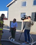 Strażak odpowiada na pytania dziennikarki, obok kamera wraz z kamerzystą, na tle budynku szkoleniowego do ćwiczeń gaśniczych