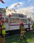 Grupa strażaków wokół zabytkowego tramwaju z śladami pożaru i unoszącym się dymem, po prawej policjant, w tle wóz strażacki