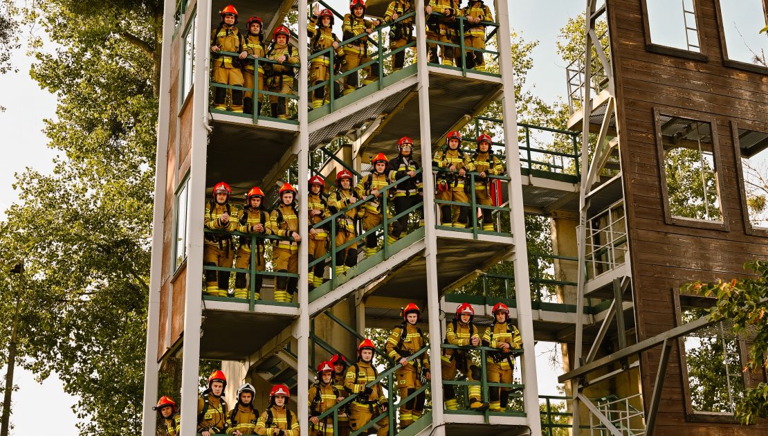 Budynek do ćwiczeń wysokościowych, na schodach stoi grupa ok. czterdziestu strażaków opierających się o barierki.