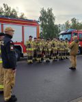 Zastępca Komendanta przemawia do grupy strażaków w dwuszeregu, po lewej stronie czterech strażaków w szeregu, na tle dwóch wozów strażackich.