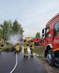 Na drodze dwóch strażaków operujących prądem wody z węża tłocznego, po prawej instruktor, po lewej dwa wozy strażackie, w tle wagon pociągu i sala gimnastyczna