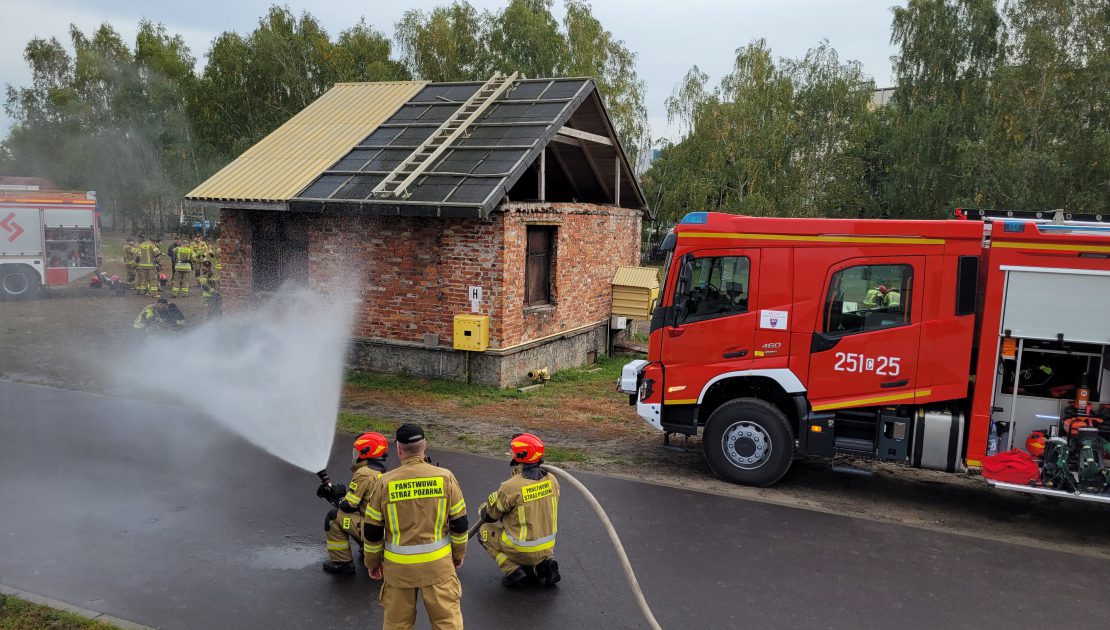 Dwóch strażaków w stroju bojowym operuje prądem wody na drodze, za nimi trzeci strażak. W tle grupa strażaków stojąca za budynkiem i wóz strażacki