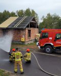 Dwóch strażaków w stroju bojowym operuje prądem wody na drodze, za nimi trzeci strażak. W tle grupa strażaków stojąca za budynkiem i wóz strażacki