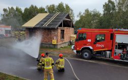 Dwóch strażaków w stroju bojowym operuje prądem wody na drodze, za nimi trzeci strażak. W tle grupa strażaków stojąca za budynkiem i wóz strażacki