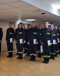 Grupa strażaków stojąca na baczność z dyplomami w dłoniach, po lewej stronie kierownik kursu w mundurze galowym.