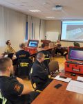 Prezentacja urządzenia ACO-Streamer, udzielana grupie strażaków siedzących w świetlicy, na slajdzie obraz z kamery drona.