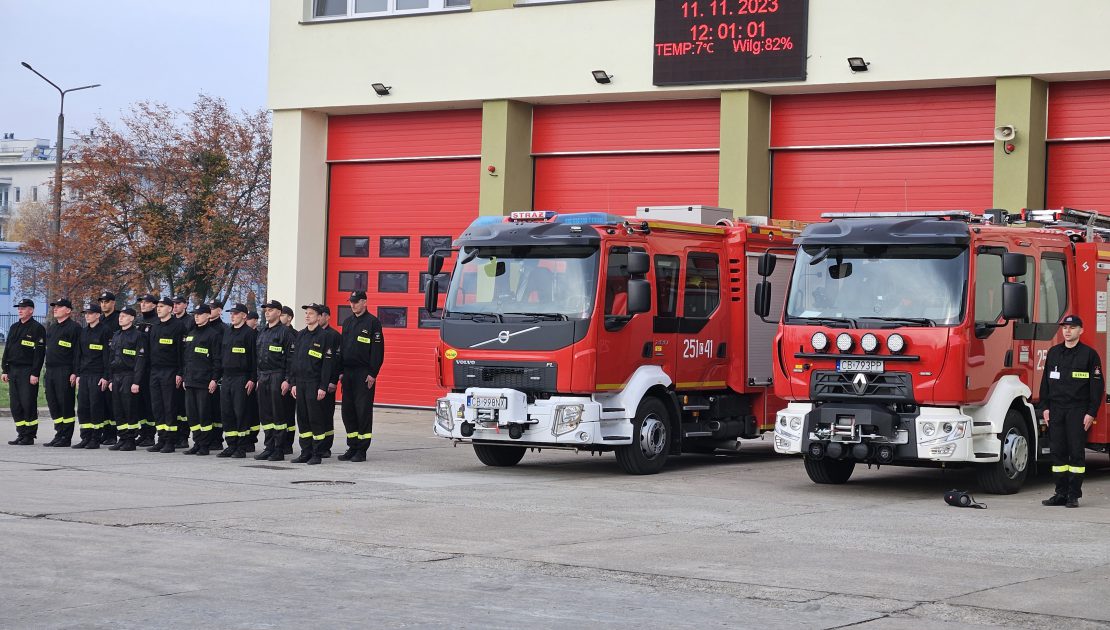 Grupa strażaków stojąca na baczność w dwuszeregu, obok dwóch wozów strażackich przed garażem bojowym JRG, dnia 11.11.2023 o godzine dwunastej.