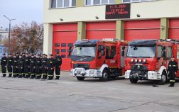 Grupa strażaków stojąca na baczność w dwuszeregu, obok dwóch wozów strażackich przed garażem bojowym JRG, dnia 11.11.2023 o godzine dwunastej.