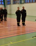 Komendant stoi przed mikrofonem, przed nim dwóch strażaków składających przysięgę, za nimi grupa strażaków i cywili na sali gimnastycznej