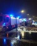 Dwa uszkodzone samochody pomiędzy dwoma wozami strażackimi w nocy, przy samochodzie grupa strażaków, koło samochodów gaśnica