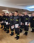 Grupa strażaków stojąca na baczność w dłoniach dyplomy ukończenia szkolenia.