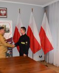 Komendant podaje rękę strażakowi, w ręku trzyma przysięgę, w tle trzy flagi Polski i baner szkoły.