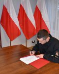Strażak podpisuje dokumenty przy stole, w tle baner szkoły oraz trzy flagi Polski