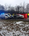 Na drodze dwa samochody po zderzeniu czołowym, wokół stoją strażacy oraz policjanci. Za samochodami stoją inne samochody, karetka pogotowia oraz wóz strażacki