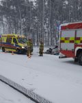 Droga pokryta śniegiem, na poboczu stoją dwa wozy strażackie a pomiędzy nimi karetka pogotowia. Pojazdy odgrodzone pachołkami drogowymi. W rowie za wozami samochód osobowy odwrócony kołami do góry.