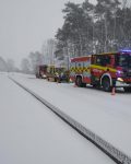 Droga pokryta śniegiem, na poboczu stoją dwa wozy strażackie a pomiędzy nimi karetka pogotowia. Pojazdy odgrodzone pachołkami drogowymi.
