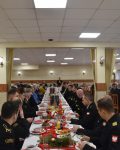 Grupa strażaków w mundurach galowych oraz cywile, siedzą przy nakrytych stołach, w oddali przemawia Komendant Szkoły Podoficerskiej.
