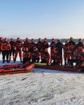 Grupa strażaków w suchych skafandrach na lodzie, pozują do zdjęcia grupowego, przed nimi ponton oraz sanie lodowe