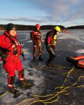 Trzech strażaków w suchych skafandrach i kaskach na lodzie, jeden z nich podpięty liną do pontonu