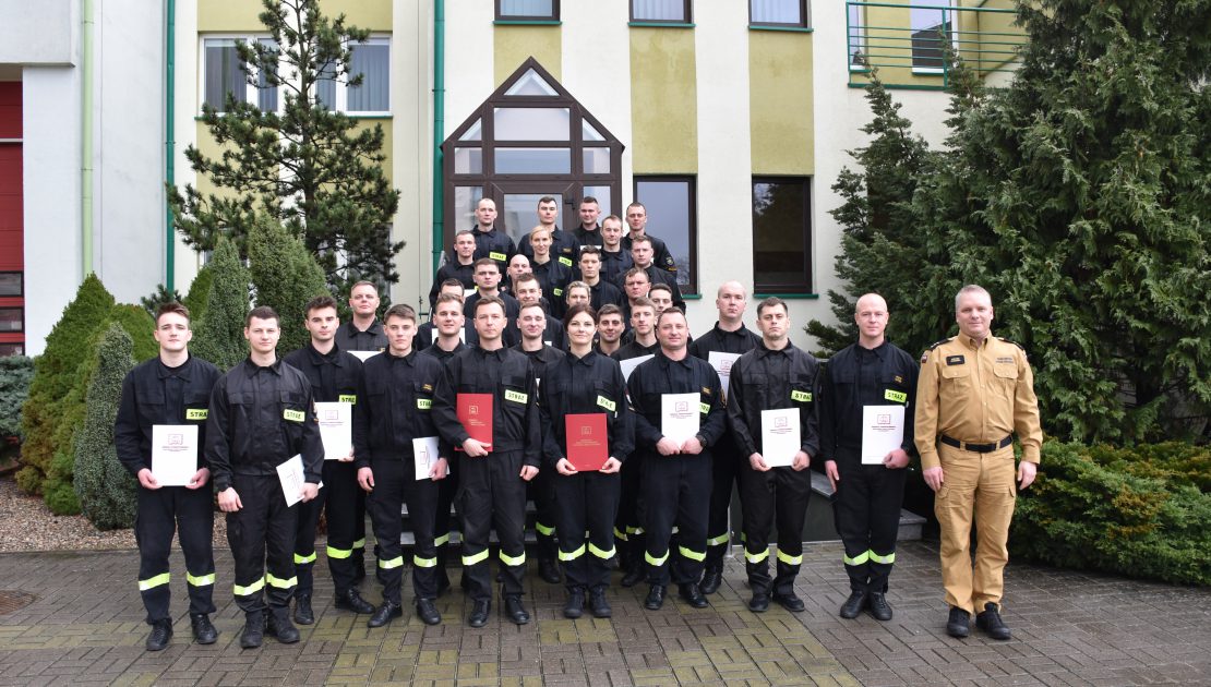 Grupa strażaków pozująca do zdjęcia na zakończenie kursu w dłoniach trzymają dyplomy, obok kierownika kursu.