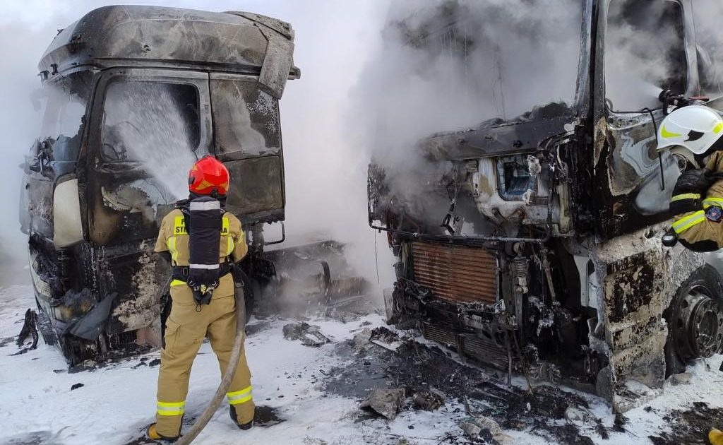 Dwa spalone samochody ciężarowe, pojazdy oraz ziemia pokryte pianą, strażak polewa kabinę jednego z samochodów pianą.