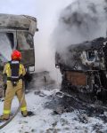 Dwa spalone samochody ciężarowe, pojazdy oraz ziemia pokryte pianą, strażak polewa kabinę jednego z samochodów pianą.
