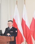 Komendant Szkoły Podoficerskiej przy mównicy, na mównicy laptop oraz mikrofon, na drugim planie znajdują się trzy flagi Polski w rzędzie.