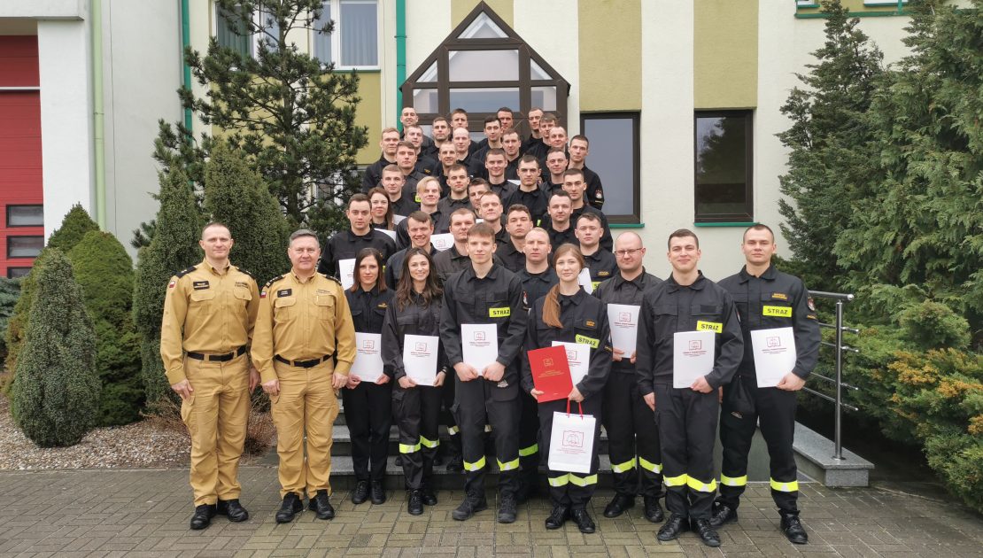 Grupa strażaków wraz z komendantem i szefem kursu, pozuje do zdjęcia na zakończenie kursu na schodach bud. administracyjnego, trzymają w dłoniach dyplomy oraz upominki.