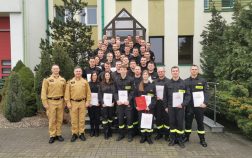 Grupa strażaków wraz z komendantem i szefem kursu, pozuje do zdjęcia na zakończenie kursu na schodach bud. administracyjnego, trzymają w dłoniach dyplomy oraz upominki.