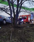 Uszkodzony samochód na wzniesieniu koło drzewa, na drugim planie wóz strażacki