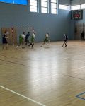 Grupa osób gra w piłkę nożną na sali gimnastycznej. Dwie drużyny walczą o piłkę w polu karnym.