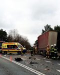 Ciężarówka z naczepą znajduje się poza drogą na której widać odłamki, na około której stoją strażacy i policjant, na drodze stoją również samochody policji, wozy strażackie oraz karetka pogotowia.