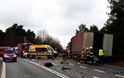 Ciężarówka z naczepą znajduje się poza drogą na której widać odłamki, na około której stoją strażacy i policjant, na drodze stoją również samochody policji, wozy strażackie oraz karetka pogotowia.