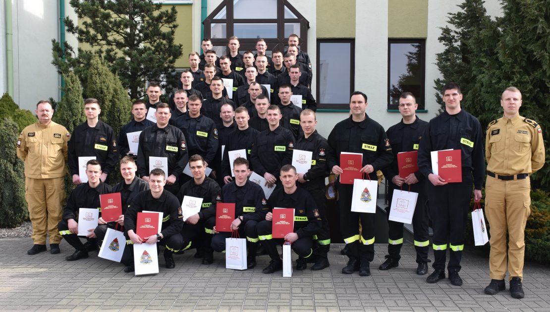 Grupa strażaków pozuje do zdjęcia grupowego wraz z dyplomami i upominkami, wraz z szefem kursu oraz komendantem.