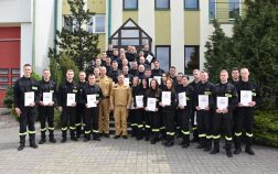 Grupa strażaków w umundurowaniu służbowym stoi przed budynkiem. Strażacy trzymają w rękach teczki z dokumentami.