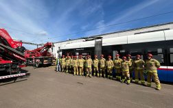 Grupa strażaków w ubraniach specjalnych stoi przed tramwajem. W tle widać samochody strażackie.