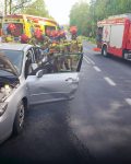 Na jezdni stoi uszkodzony samochód osobowy. Z tyłu pojazdu działa grupa strażaków. W tle dwa wozy strażackie i ambulans