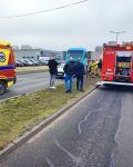 Na jezdni z lewej strony stoi ambulans, po prawej stronie wóz strażacki. W tle grupa strażaków i 3 osoby stojące przy uszkodzonych pojazdach.