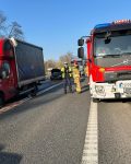 Po lewej stronie stoi uszkodzony samochód dostawczy. Po prawej stronie wóz strażacki. Z tyłu w tle widać samochody. Z przodu stoi strażak i policjant.