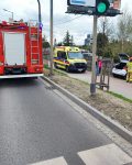 Na jezdni stoi samochód strażacki. Na chodniku ambulans. Samochód osobowy wjechał w betonowe ogrodzenie, które jest uszkodzone. Dwóch strażaków stoi przy ogrodzeniu.